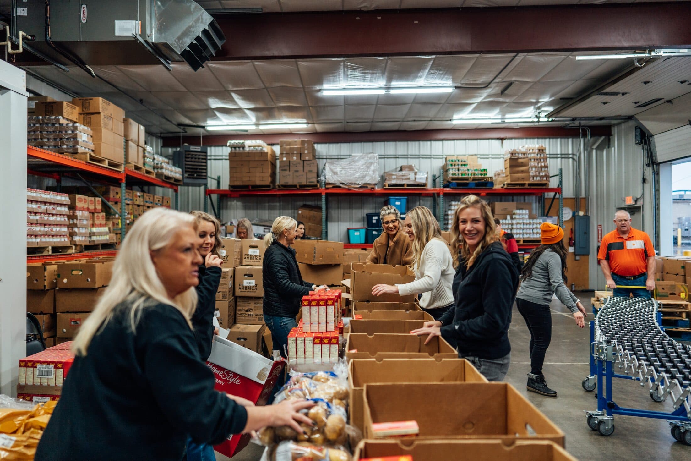 Volunteers filling cardboard boxes with groceries along a warehouse line for a holiday basket drive.