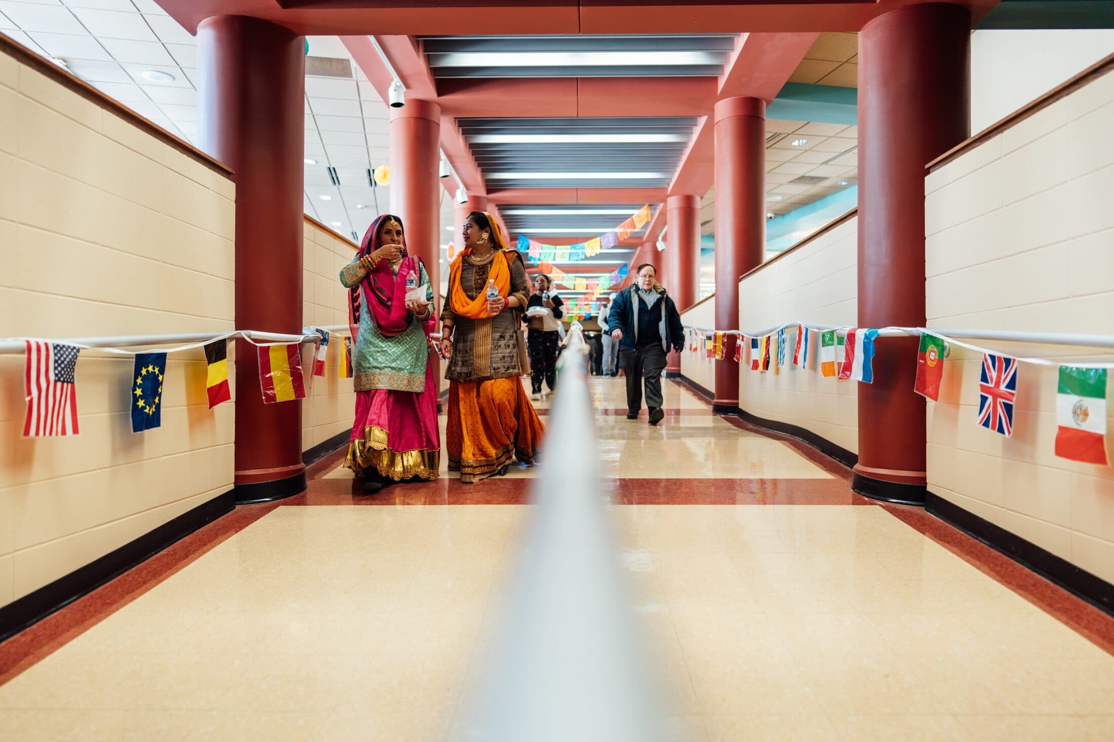A school corridor lined with the flags of many nations, students walking toward the camera in traditional dress.