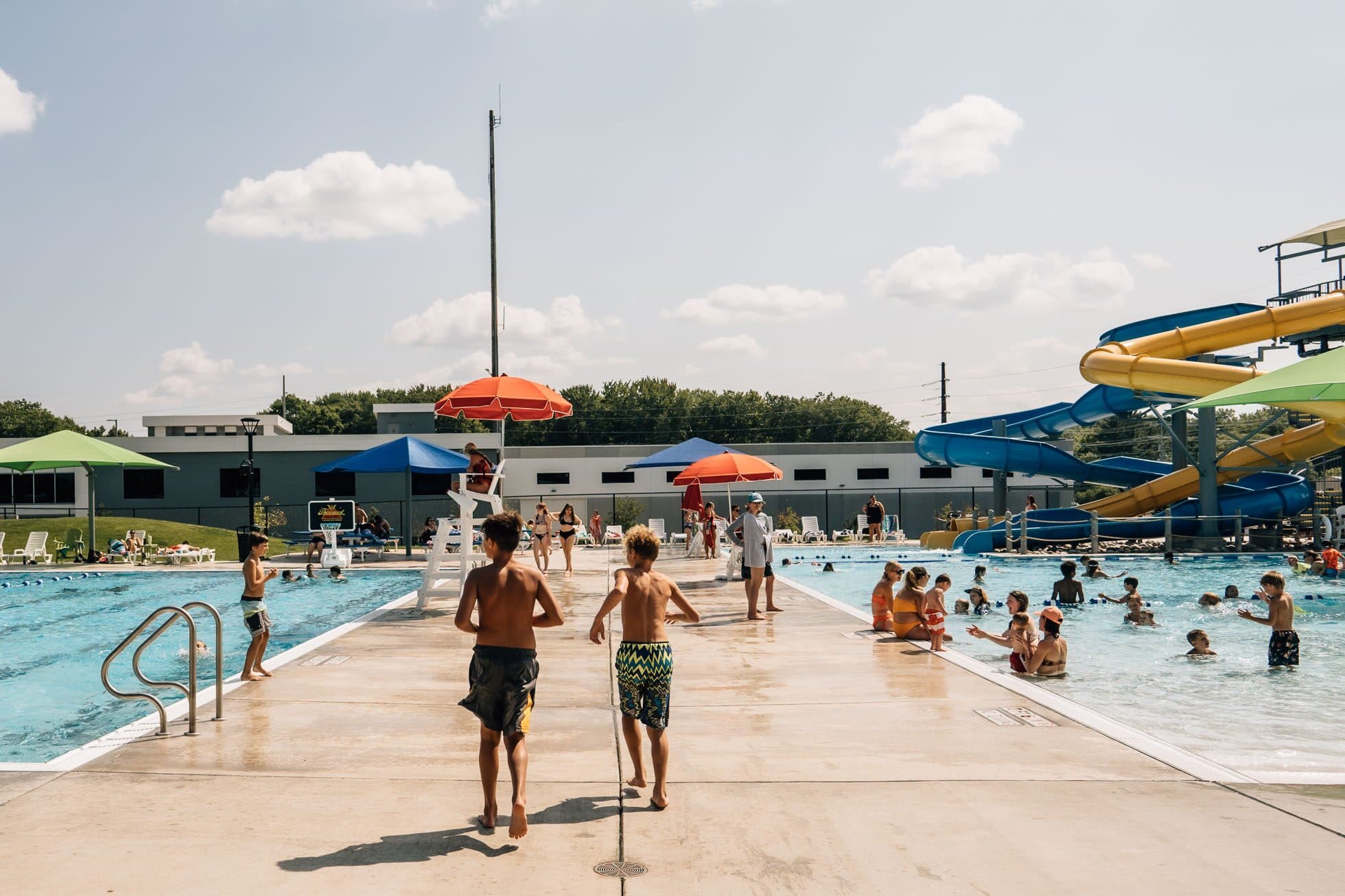 Children walking the deck of a busy municipal pool on a bright summer afternoon.